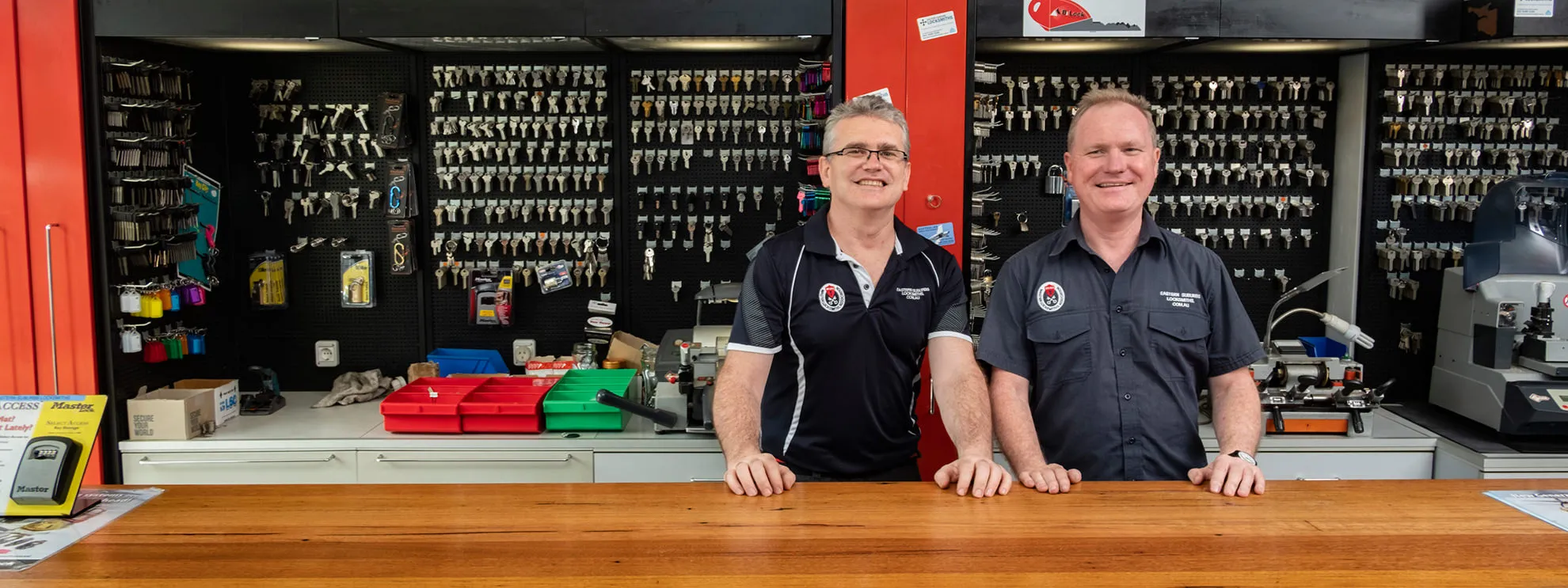 Two locksmith staff standing behind a shop counter with key blanks displayed on the wall