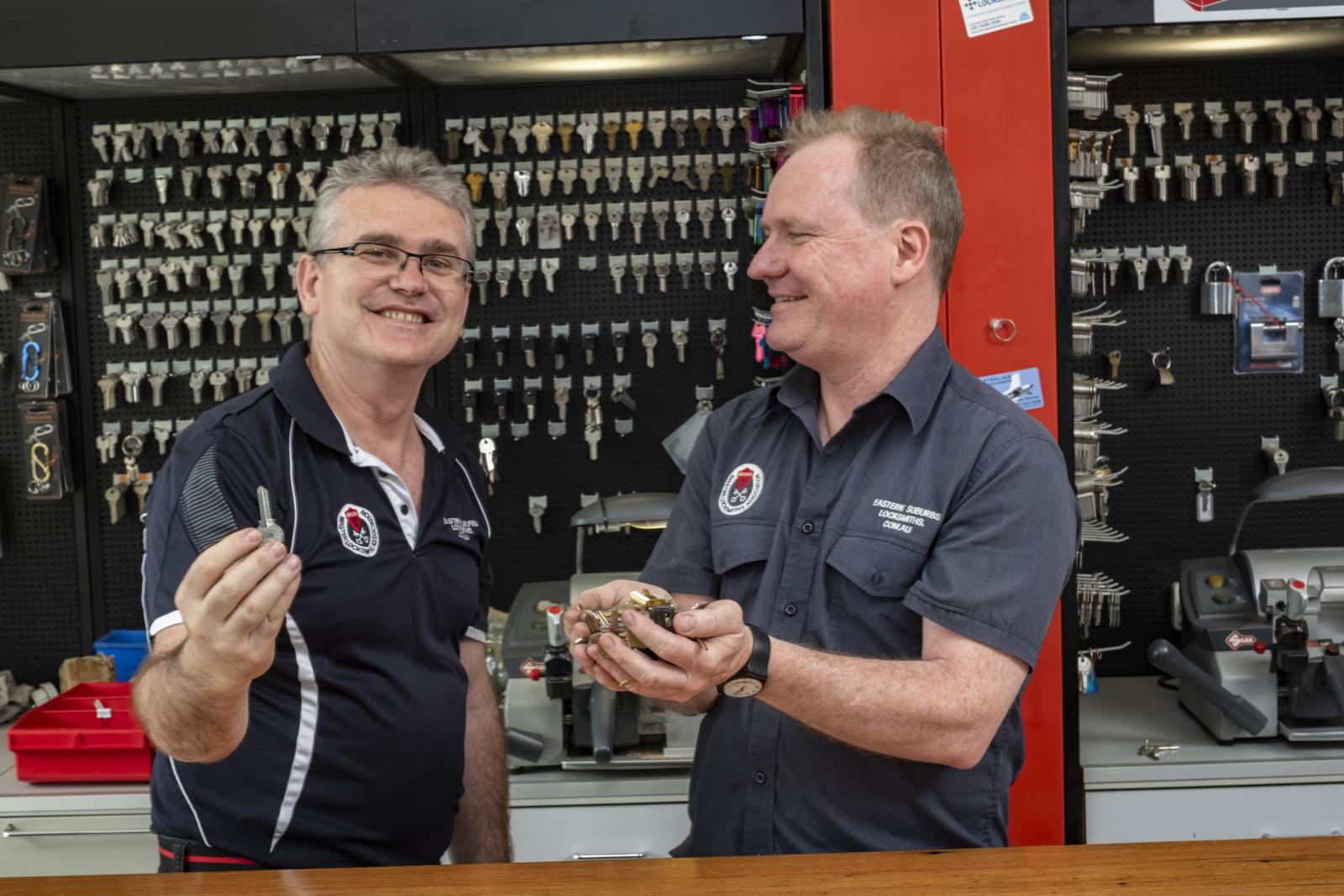 Two locksmiths holding keys in a workshop with key blanks displayed behind them
