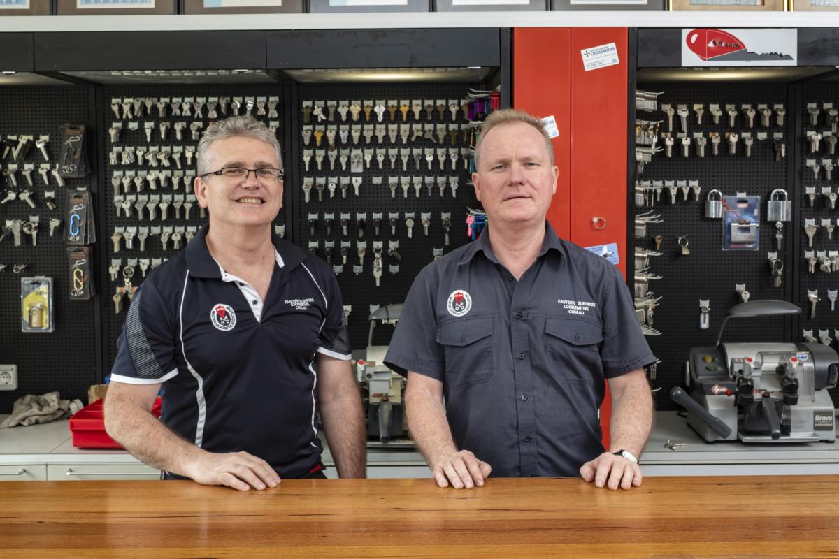 Two locksmiths standing behind a shop counter with key blanks and cutting equipment in the background