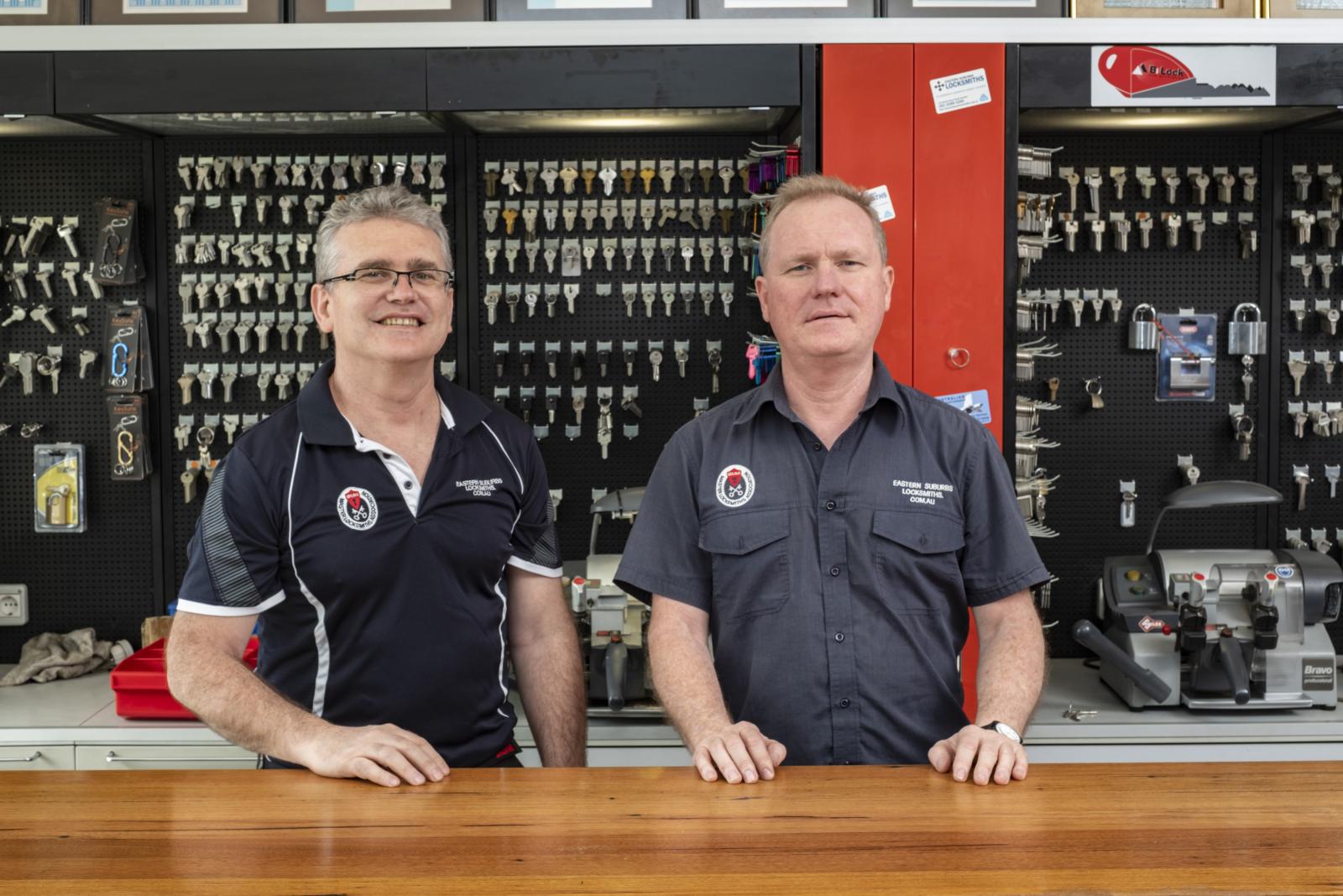 Two locksmiths standing behind a shop counter with key blanks and cutting equipment in the background
