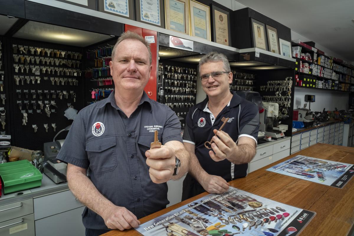Two locksmith staff holding keys at a shop counter with key boards behind them