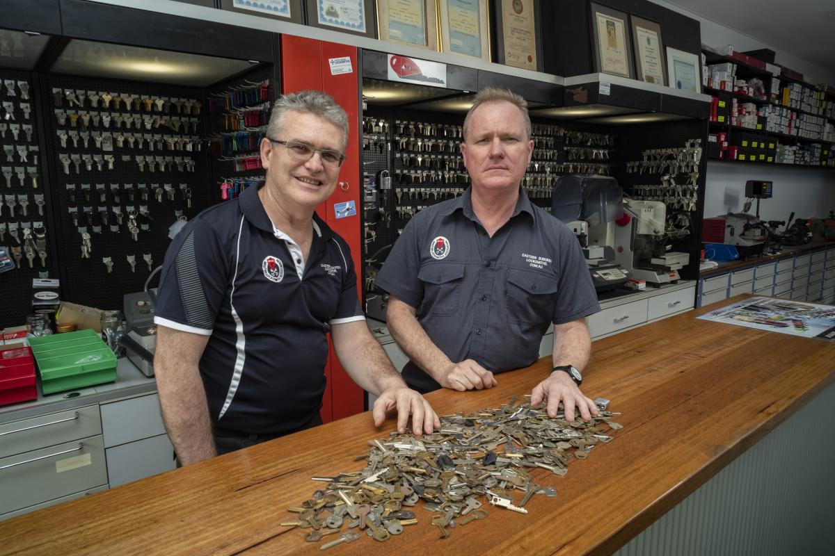 Two locksmiths standing behind a shop counter with a large pile of keys