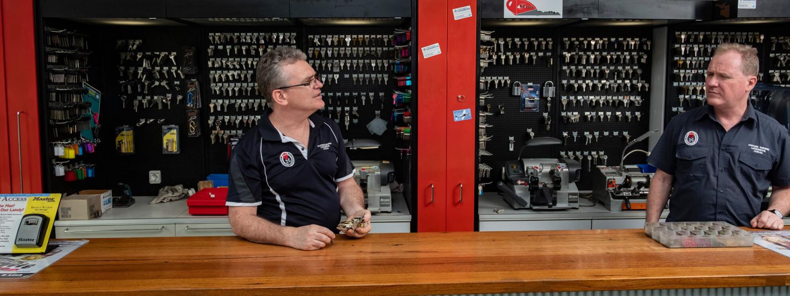Two locksmiths at a shop counter with key blanks displayed behind them